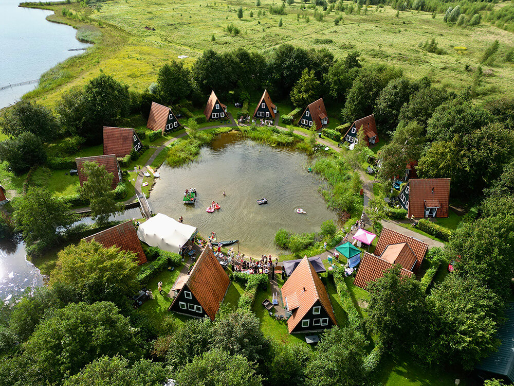 Dronefoto van een vakantiepark met puntdakhuisjes rond een vijver